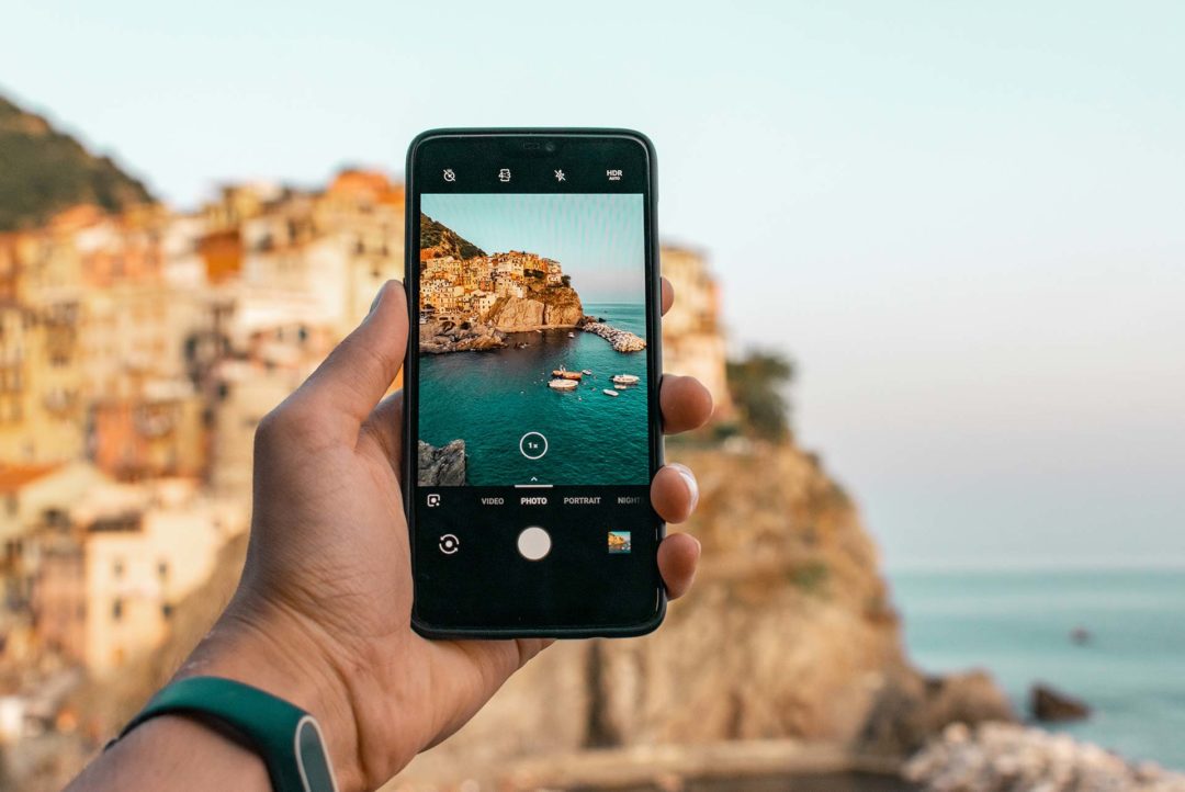 Uma mão segurando um celular que está com a câmera ligada. Na tela do aparelho pode-se ver o que estava sendo fotografado: uma pequena cidade próxima ao mar.