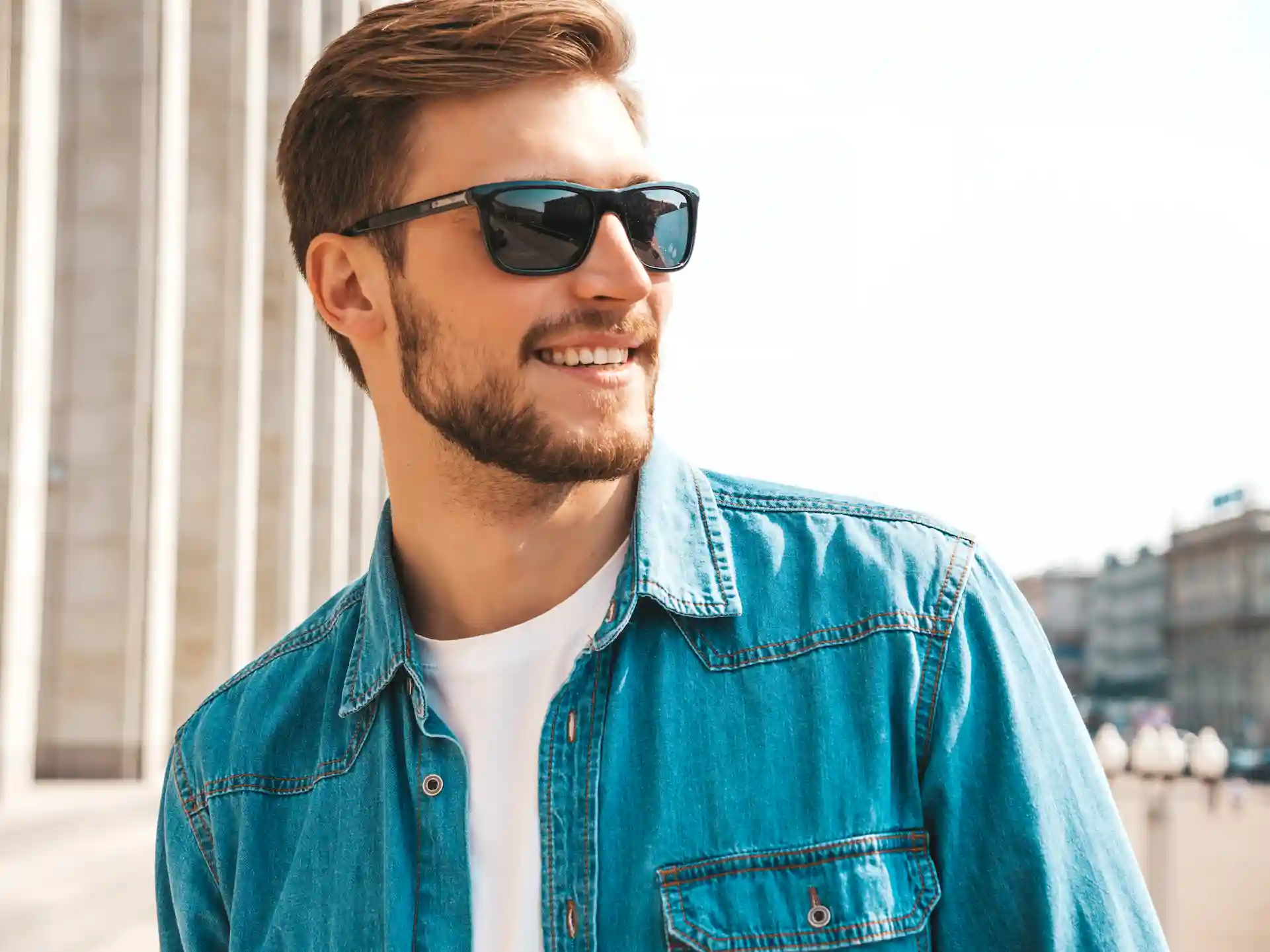 Homem utilizando camiseta branca por baixo de uma jaqueta jeans azul, e óculos de sol