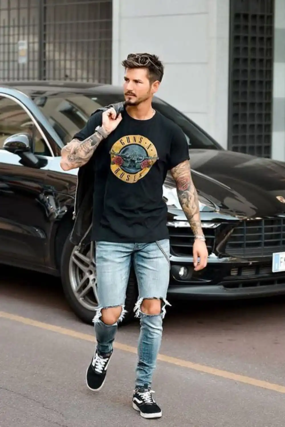 Homem utilizando camiseta preta com estampa de banda de rock, calça jeans azul rasgada e tênis.