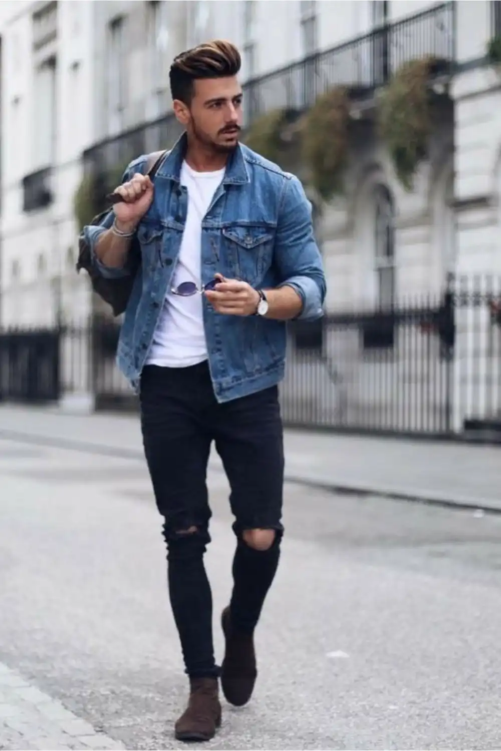 Homem utilizando camiseta branca, calça preta rasgada, jaqueta jeans azul e bota.