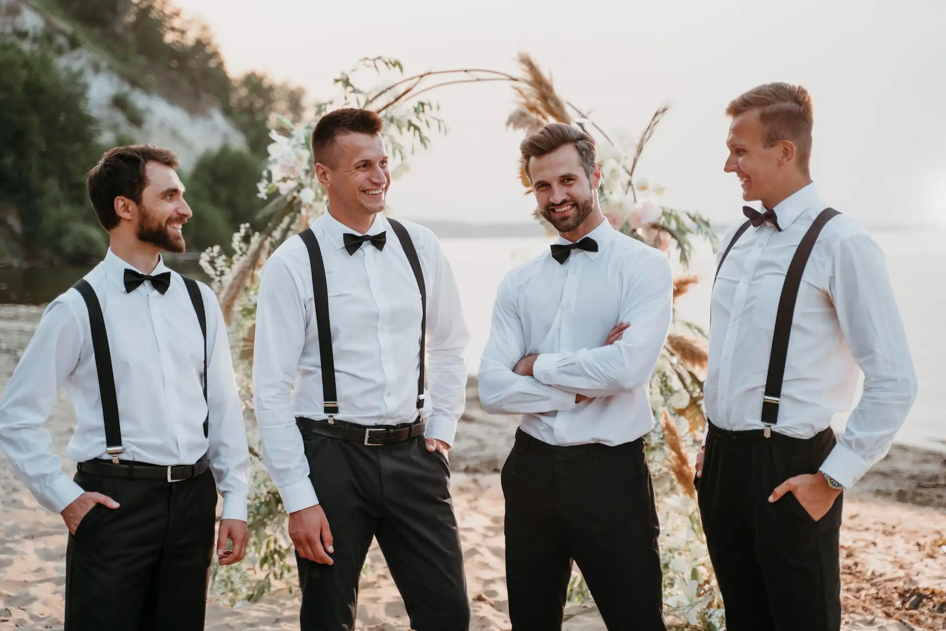 Homens em um casamento na praia utilizando peças de um traje black tie, mas sem o smoking. Esse é um típico look masculino para casamento.